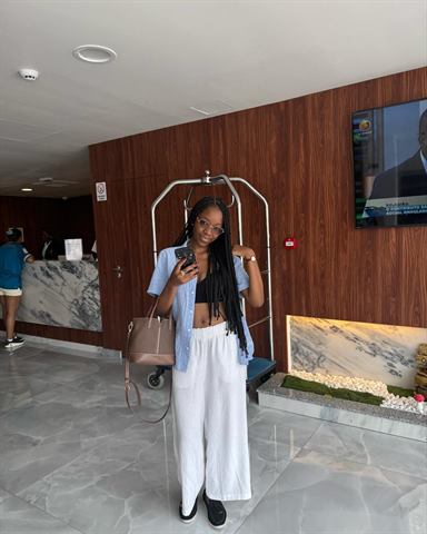 A Claudia num hall de hotel, com a cabeça inclinada, óculos, mala castanha e calças brancas largas